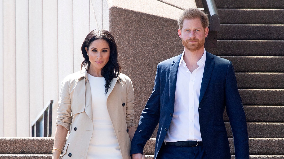 Meghan con un abrigo beige y un vestido blanco camina de la mano del príncipe Harry una chaqueta azul y una camisa blanca.