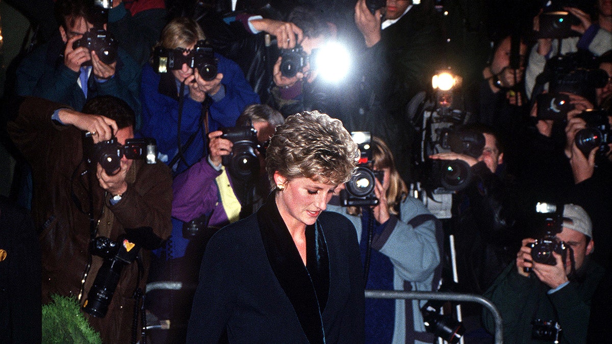 Princess Diana keeping her head down as paparazzi surround her from a metal fence.