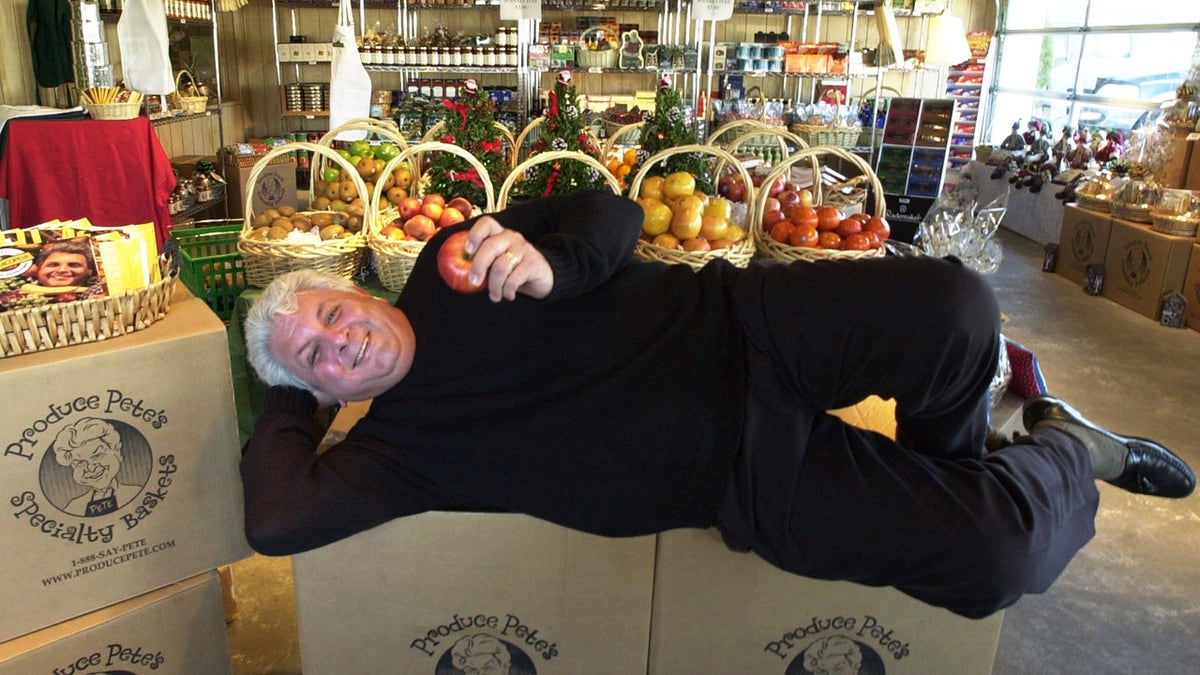 Peter "Produce Pete" Napolitano laying across boxes of his branded specialty fruit, holding apple and smiling, posing, at NJ market.