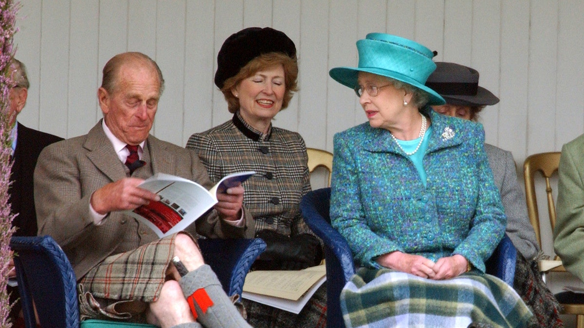 Queen Elizabeth looking at Prince Philip who is reading a book and making funny faces.