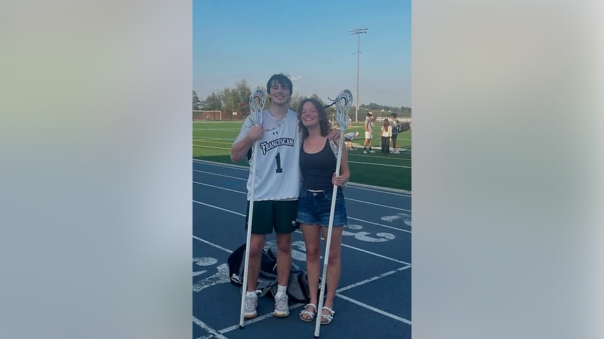 A boy and girl stand next to each other holding lacrosse sticks on a track.