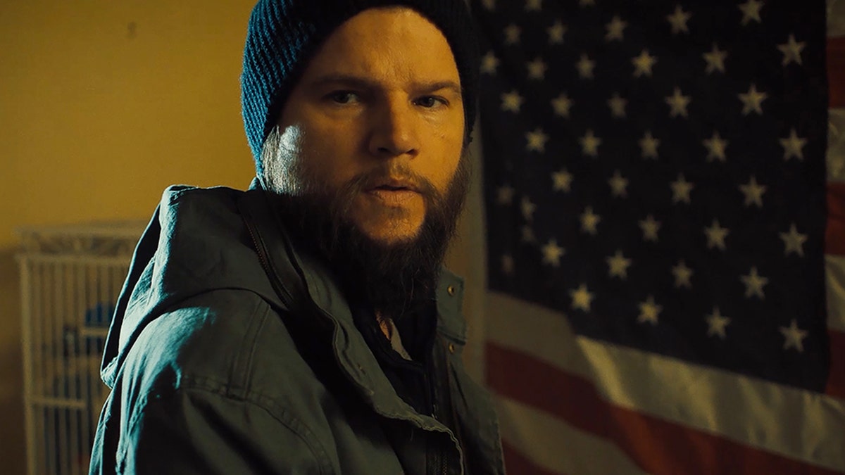 A close-up of Steven Grayhm standing in front of an American flag while acting out a scene in "Sheepdog."