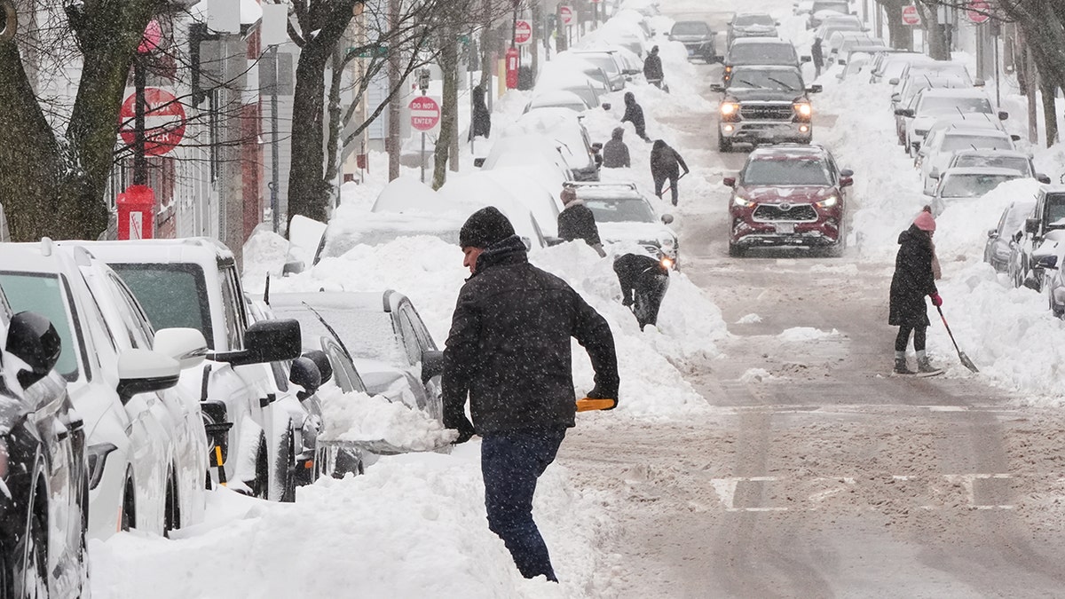 People using shovels to dig out parked cars covered in deep snow on a city street.