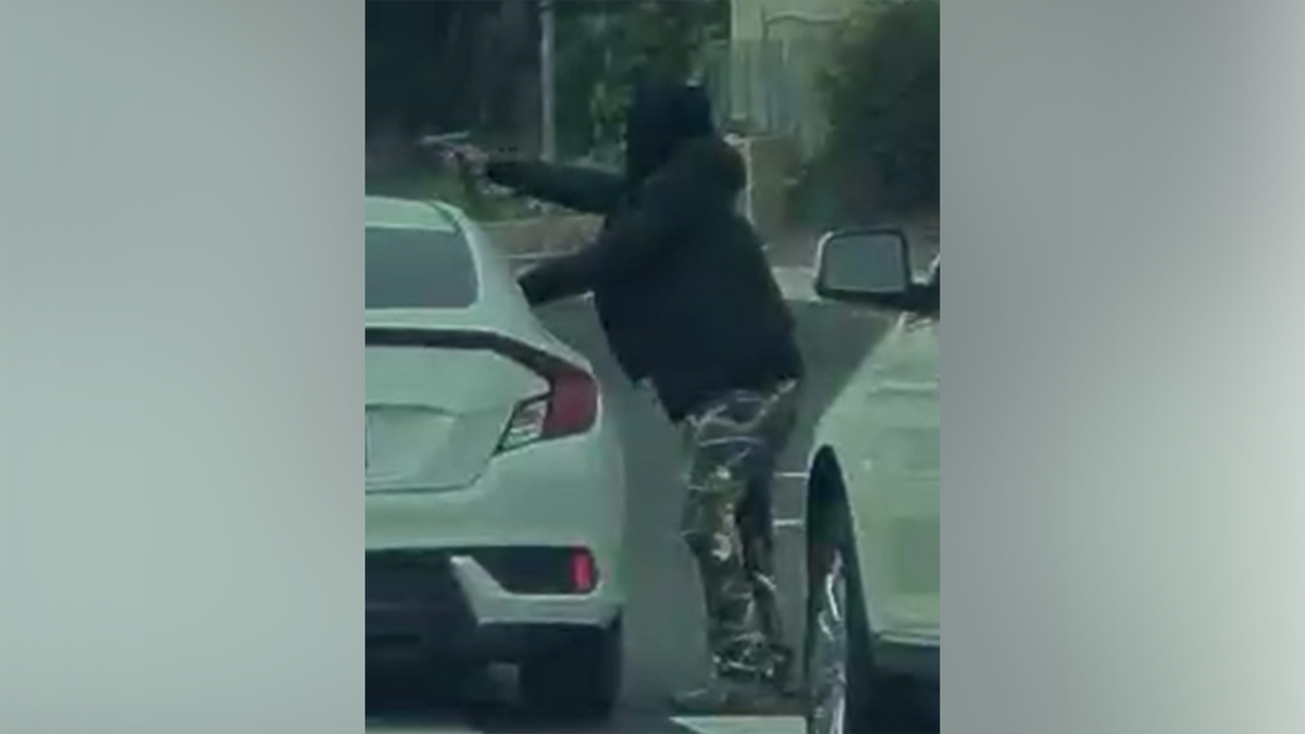 A person appears to point a handgun while standing between vehicles on a roadway