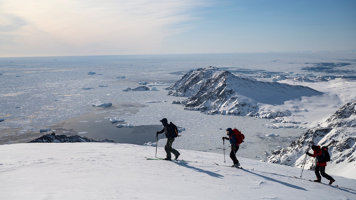 Three ski mountaineers climb a snow-covered ridge overlooking an icy Arctic coastline and frozen sea below.
