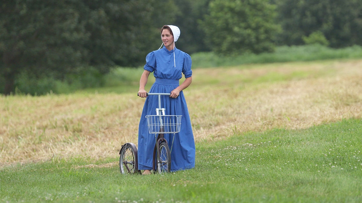 A woman wearing traditional blue Amish fashion holding a bike in a field.