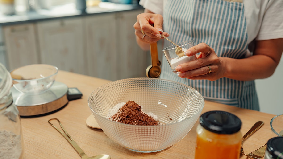 Woman putting baking powder into bowl of dry ingredients for baking recipe, seen in kitchen with apron on and scale and other ingredients around her.