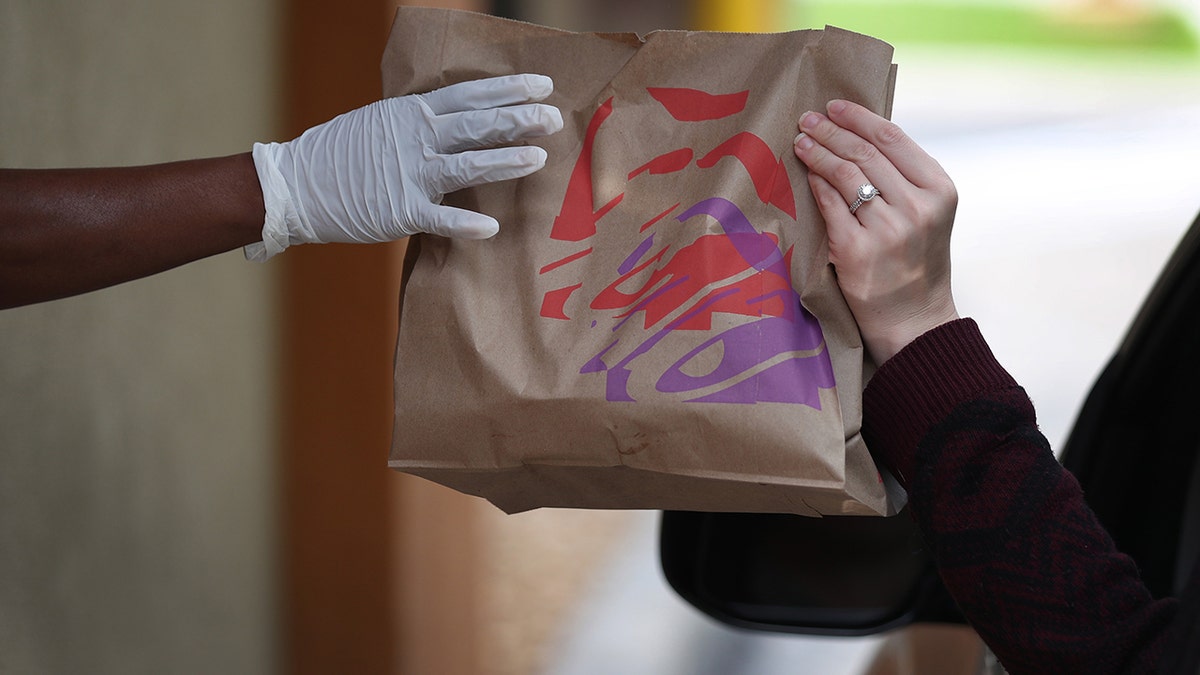 Taco Bell employee wearing glove handing bag of food through drive-thru window to customer in car.