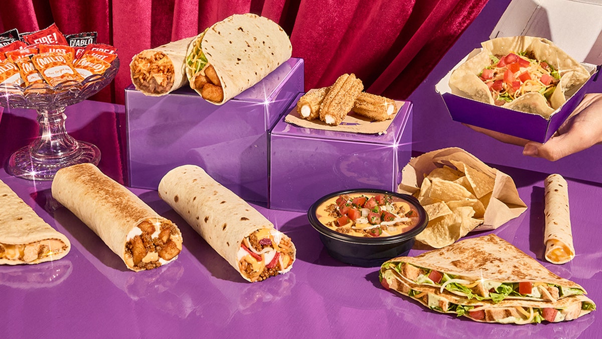 Items from Taco Bell's new Luxe Value Menu, including the Mini Taco Salad, Beefy Potato Loaded Griller, Chips & Nacho Supreme Dip, Avocado Ranch Chicken Stacker and Salted Caramel Churros. Crystal bowl filled with hot sauce packets and hand outstretched holding taco salad bowl.