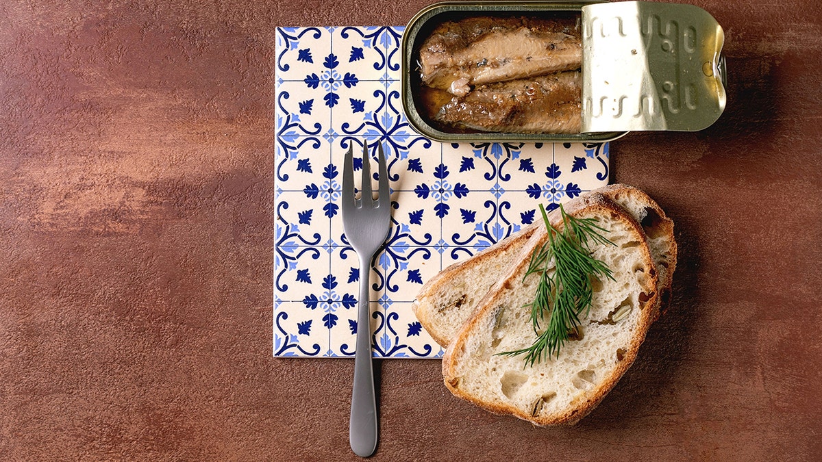 Uma lata aberta de filés de sardinha em azeite é servida com pão de trigo fresco e endro, com um garfo apoiado em azulejos de cerâmica estampados em branco e azul sobre um fundo marrom texturizado, fotografado de cima.
