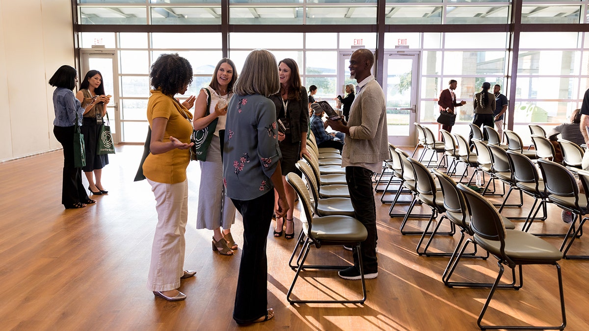 Group of professionals networking and talking inside a modern conference venue with rows of chairs and large windows.