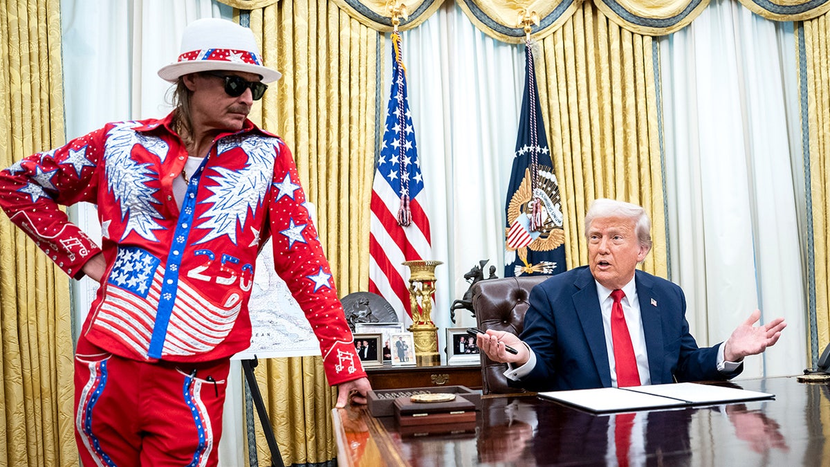 Donald Trump speaks to reporters while signing a document as Kid Rock stands beside him in the Oval Office.