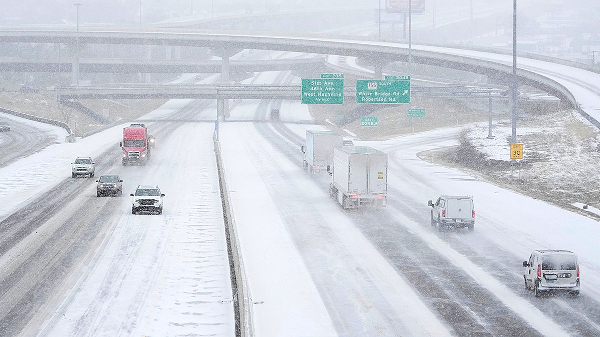 Cars and trucks drive along a highway as snow falls and roads appear slick during a winter storm.