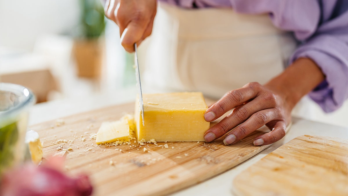 Mani che affettano un blocco di formaggio con un coltello su un tagliere di legno in una cucina.