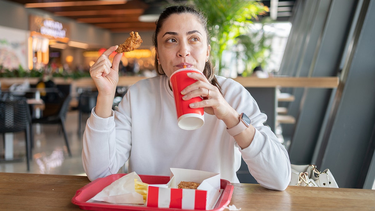 Donna che beve soda e tiene in mano un'ala di pollo fritto mentre mangia un pasto fast food in una moderna food court.
