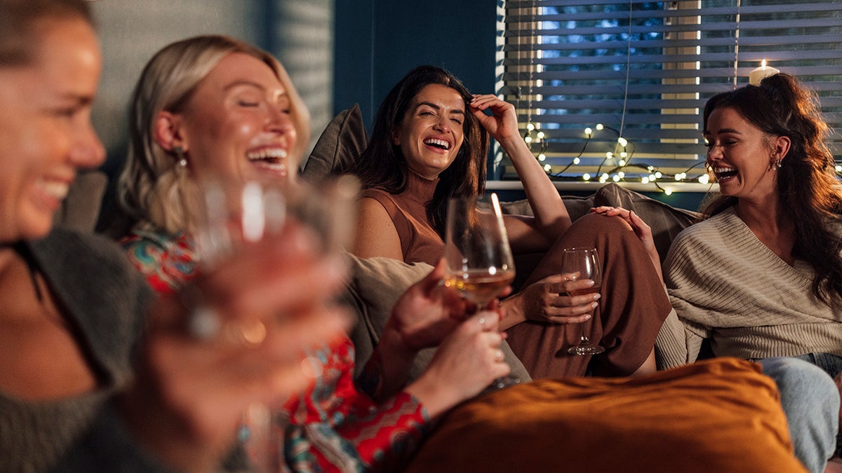 Group of women sitting at home on couch laughing drinking wine and other drinks.
