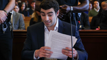 Luigi Mangione wearing a dark jacket and collared shirt while holding papers at the defense table in a Manhattan courtroom