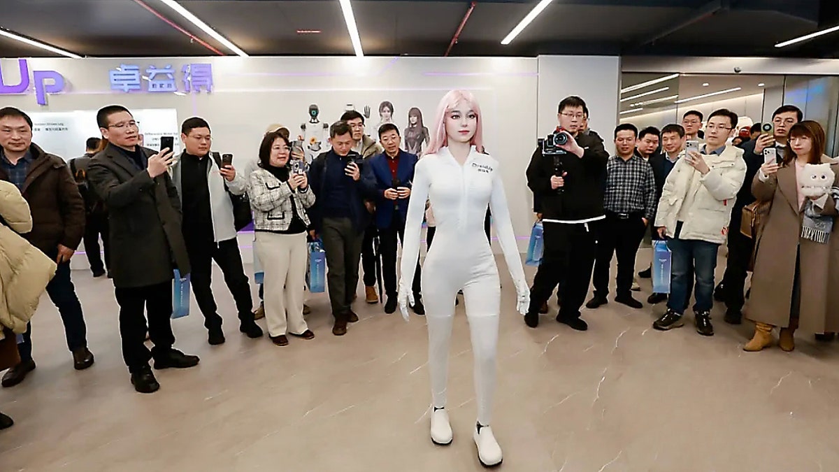 Human-like robot standing still in front of a crowd of people taking pictures.