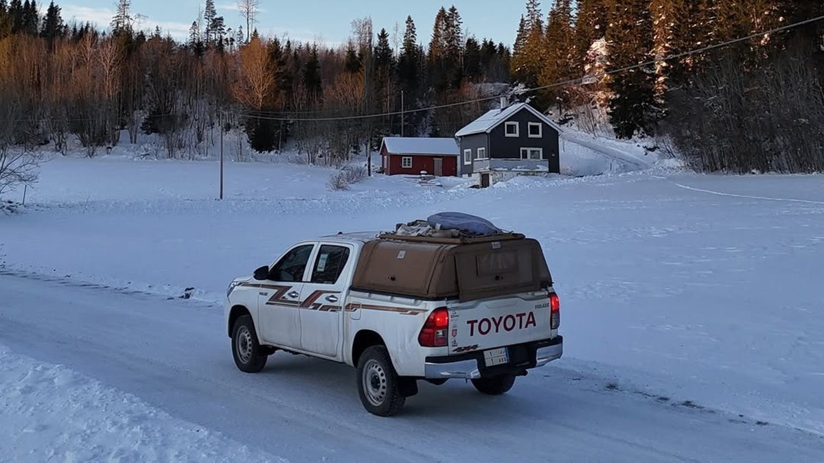 White Toyota pickup truck driving on a snowy rural road with cabins and pine forest in a winter landscape.
