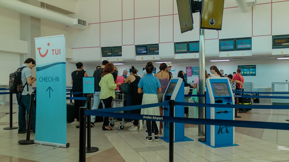 Viaggiatori in fila ai banchi del check-in all'interno dell'aeroporto di Aruba, con chioschi self-service e segnaletica aerea visibili.