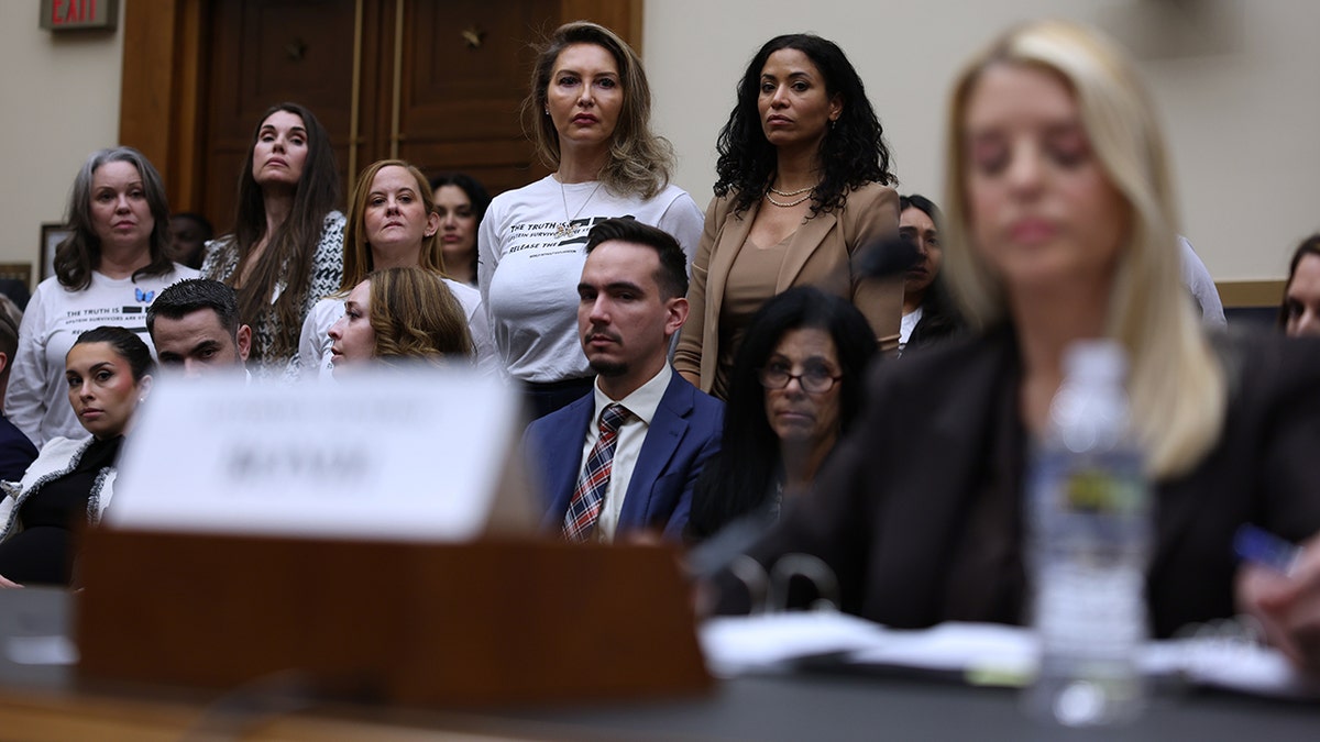 Attorney General Pam Bondi testifying at a hearing while a group of people stands to the left.