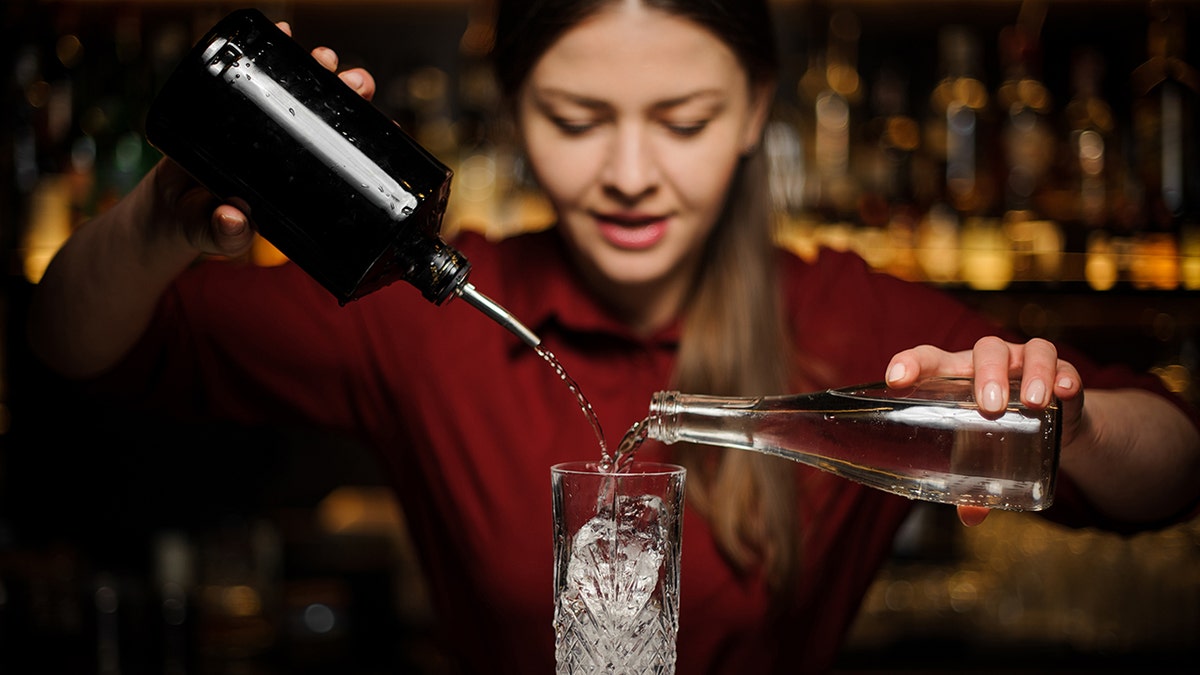 Female bartender in a red shirt behind a bar counter prepares an alcoholic cocktail Caipirinha, pouring from two bottles into a crystal glass with ice