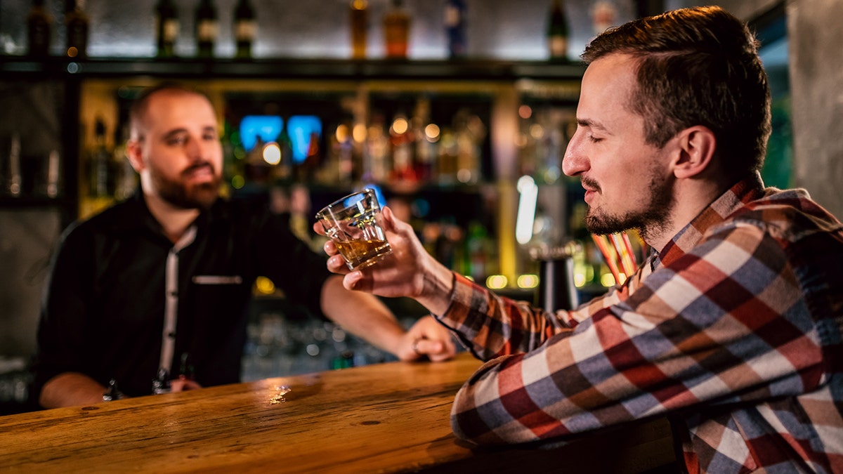 Man eyeing class if liquor in his hand at bar while talking with bartender.