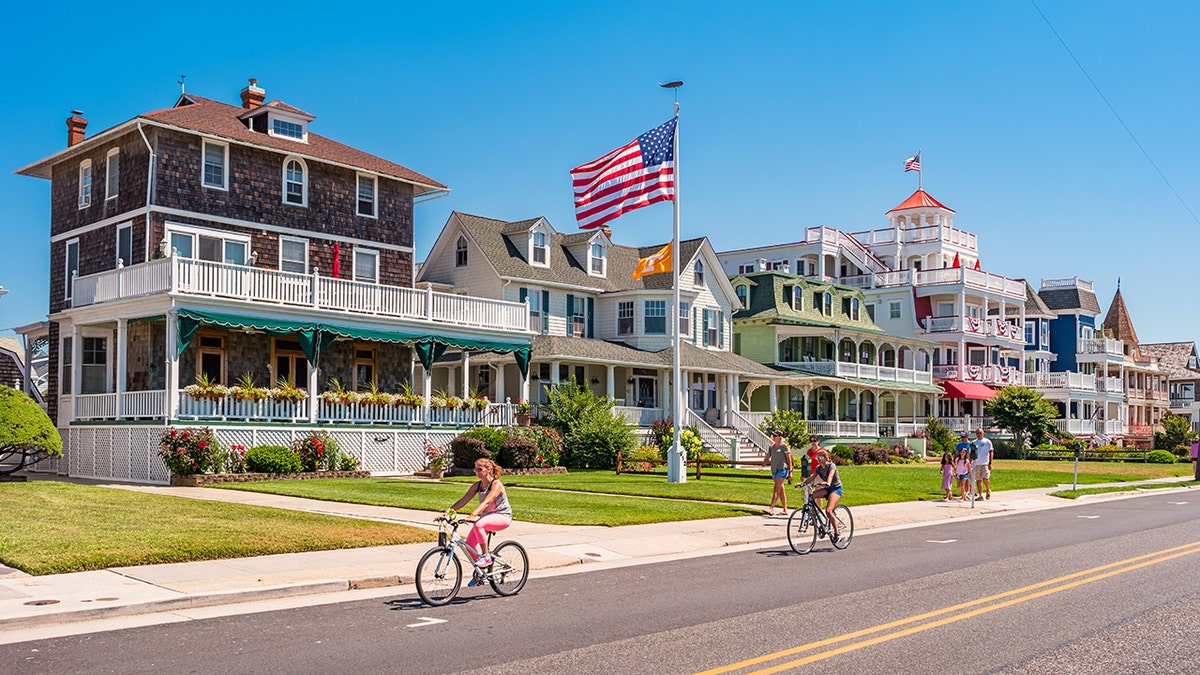 Victorian-style seaside homes with American flags along a sunny coastal street where people ride bicycles.