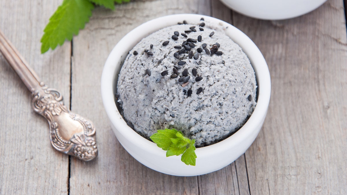 Bowl of gray sesame ice cream in white bowl, garnished with black sesame seeds and mint, with antique spoon alongside on wood table.