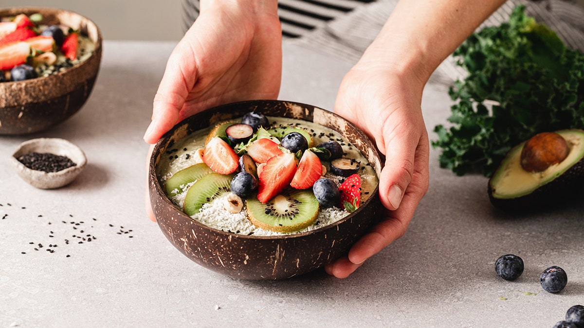 Woman's hands holding healthy smoothie bowl with different fruits in kitchen.