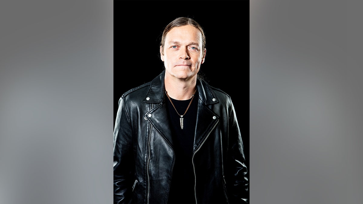 Brad Arnold wearing a leather jacket in front of a black backdrop.