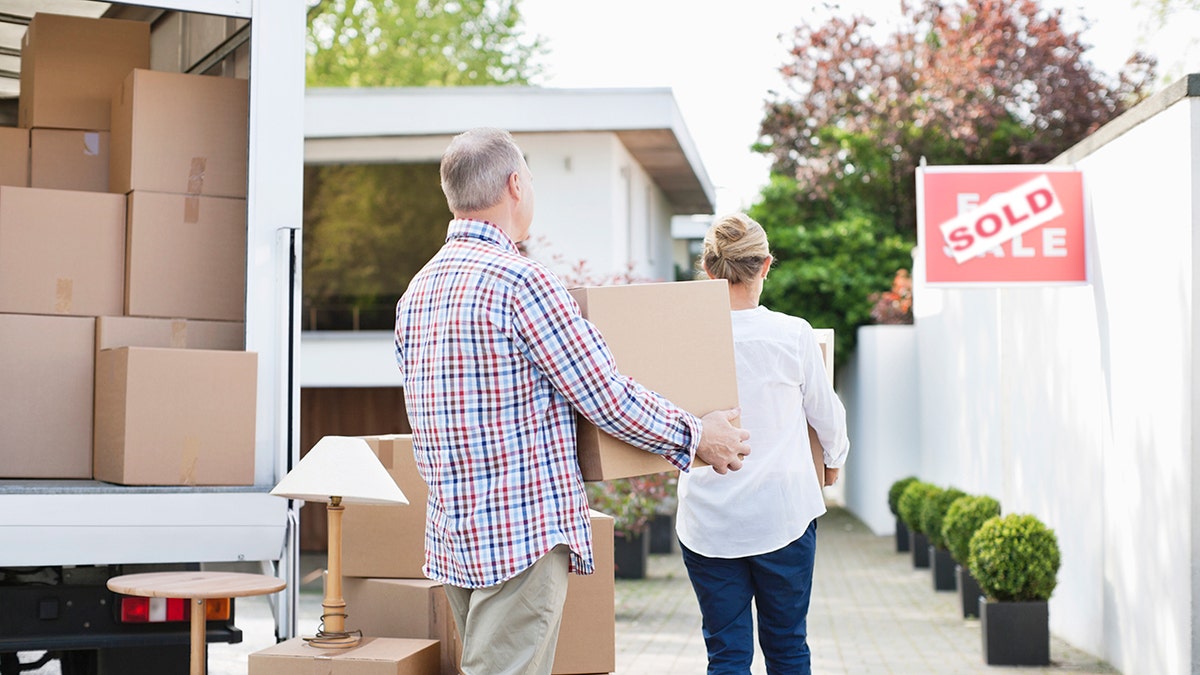 Couple carrying moving boxes into a new home with a sold real estate sign visible in the yard.