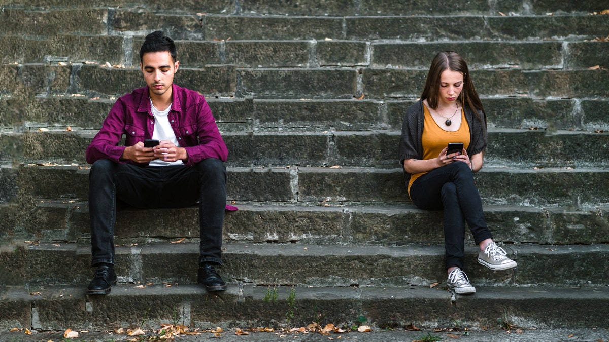 Young man and woman sitting on steps texting with big space in between.