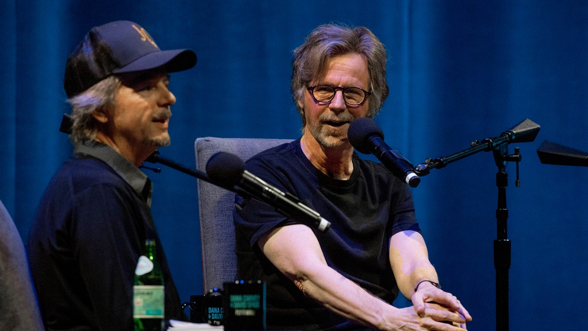 David Spade and Dana Carvey seated at microphones on a stage during an episode of "Fly on the Wall."