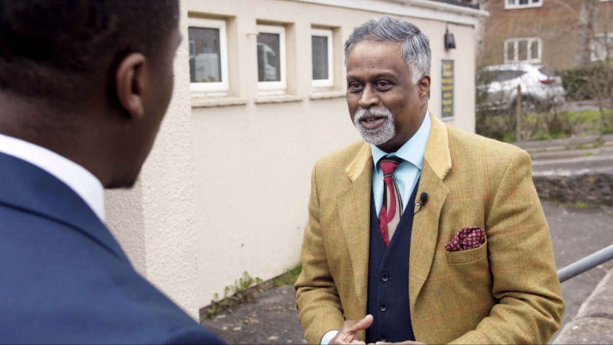 Dia Moodley speaking to his lawyer while wearing a tan blazer and a patterned tie.