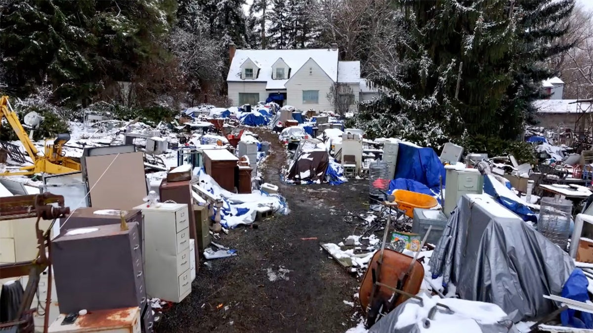 Snow-covered yard filled with scattered furniture, appliances, and tarps leading up to a white house surrounded by tall trees.