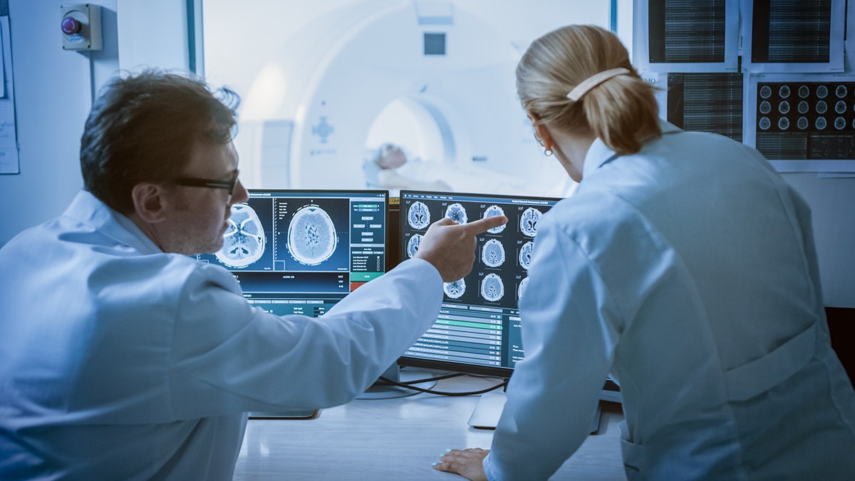 Doctors analyzing brain MRI scans on computer monitors in a hospital radiology control room.