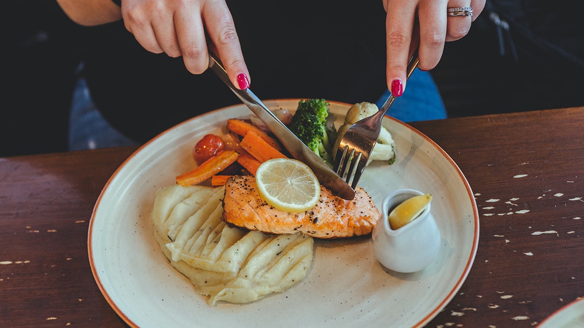 Woman's hands seen cutting into fish alongside plate of mashed potatoes and veggies at restaurant.