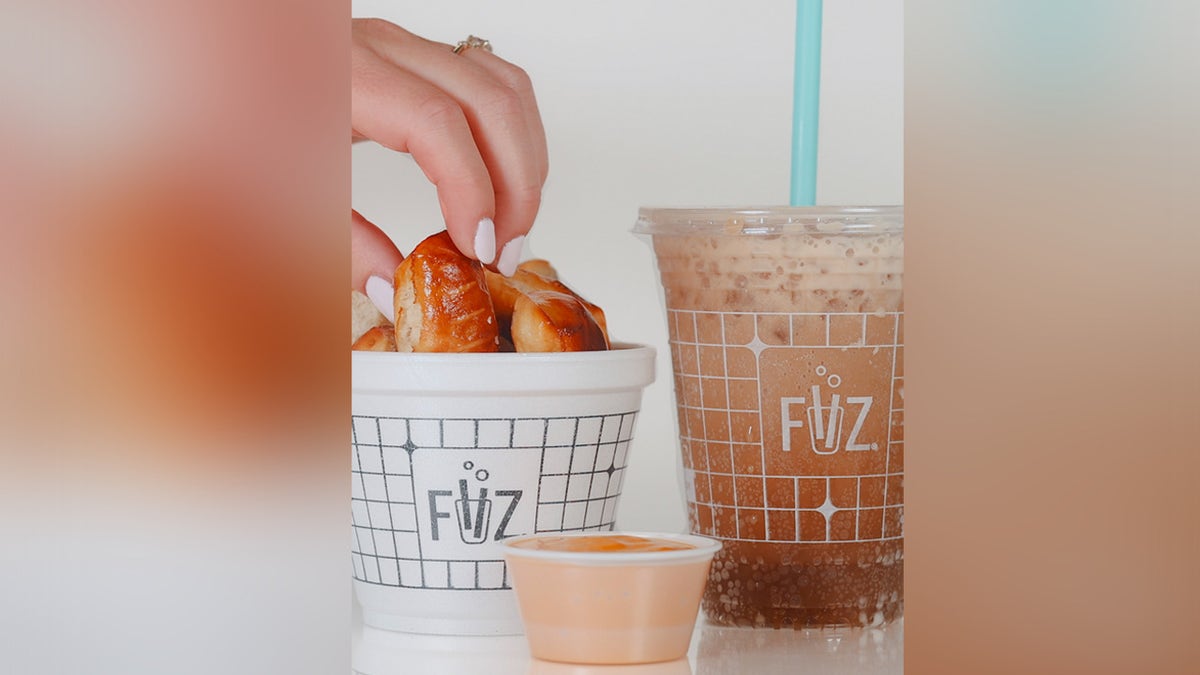 Hand grabbing pretzel bite next to "dirty soda" beverage and little cup of dipping cause.