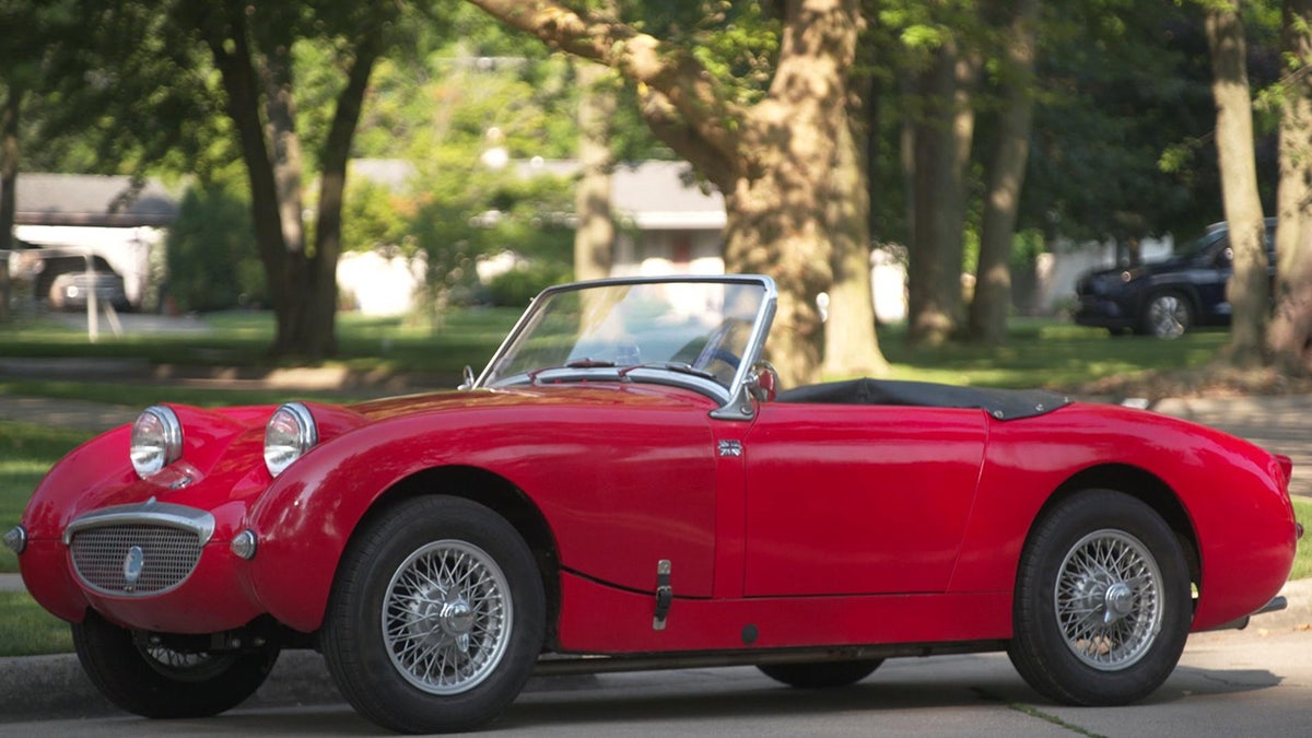 Red vintage convertible sports car parked on a residential street with trees and houses in the background.