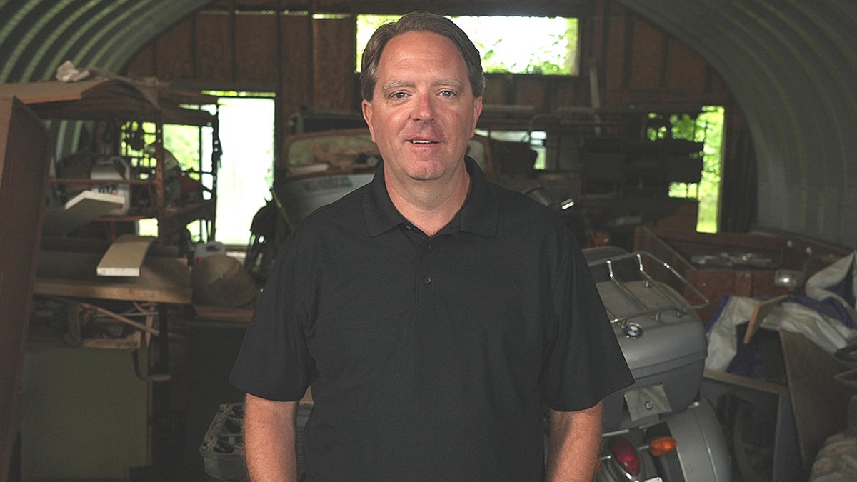 Matt Paxton standing inside a garage or storage shed filled with tools, equipment, and a motorcycle behind him.