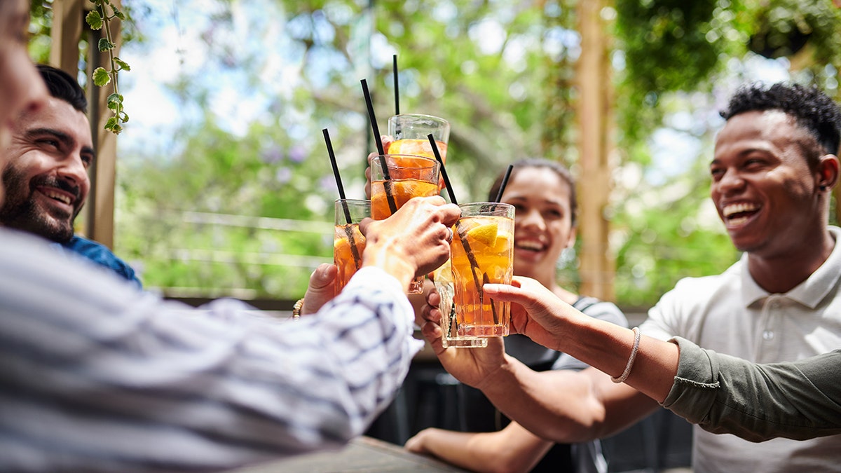 Gruppo di amici che tintinnano cocktail con fette d'arancia in un bar con patio all'aperto circondato dal verde.