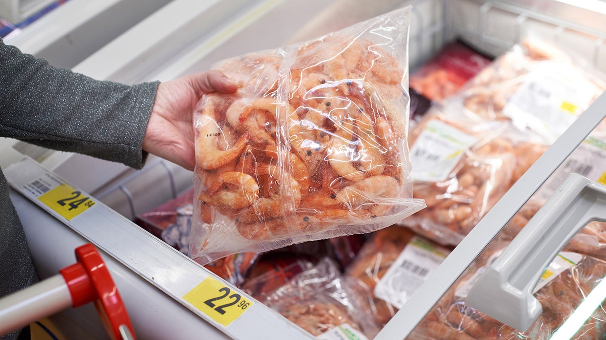 Woman grabbing frozen shrimp bag from freezer at grocery store.