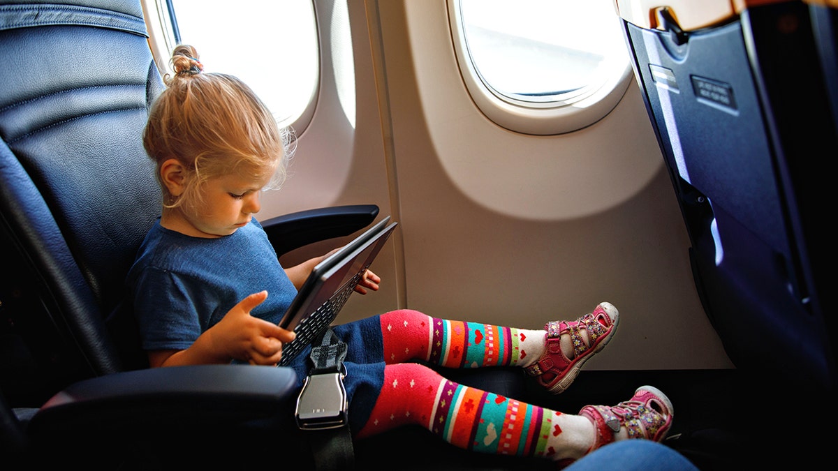 Toddler girl using tablet while seated by airplane window on a commercial flight.