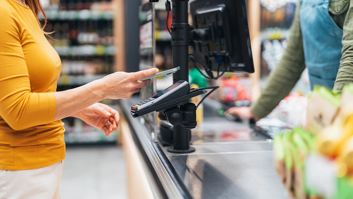 Woman uses phone for tap to pay at grocery store, cashier seen on other side of register