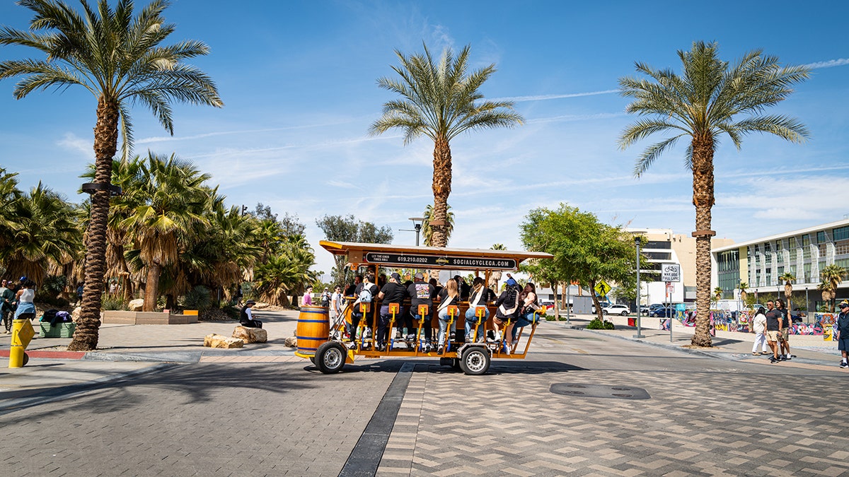 Group of people riding a pedal-powered party bike along a palm-lined city street on a sunny day.