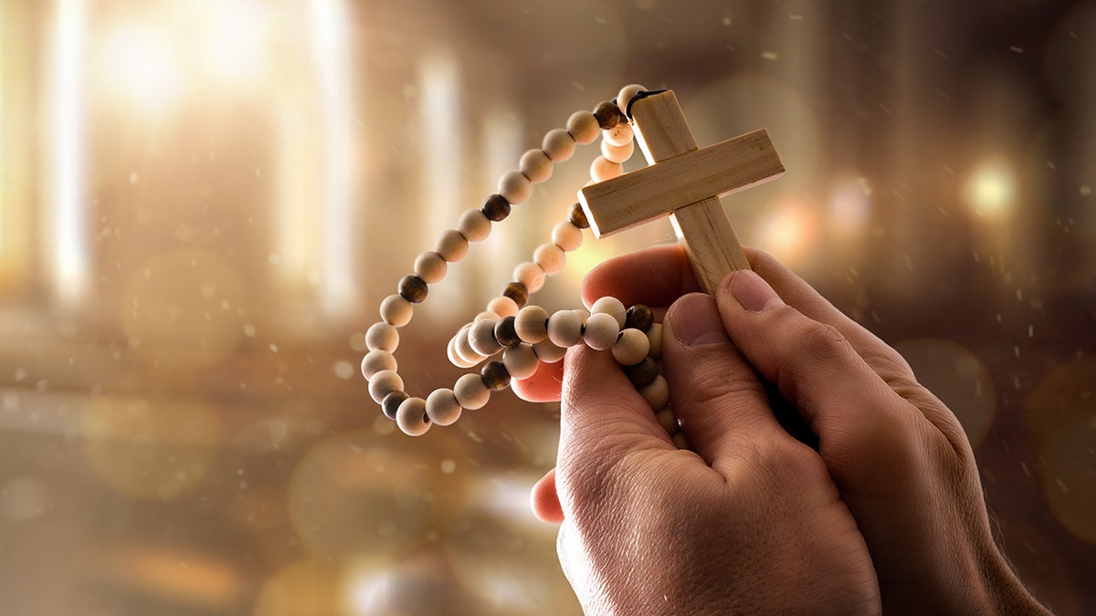 Man holding religious cross with wooden beads in a large religious room with large windows letting in sunlight and with a spiritual atmosphere