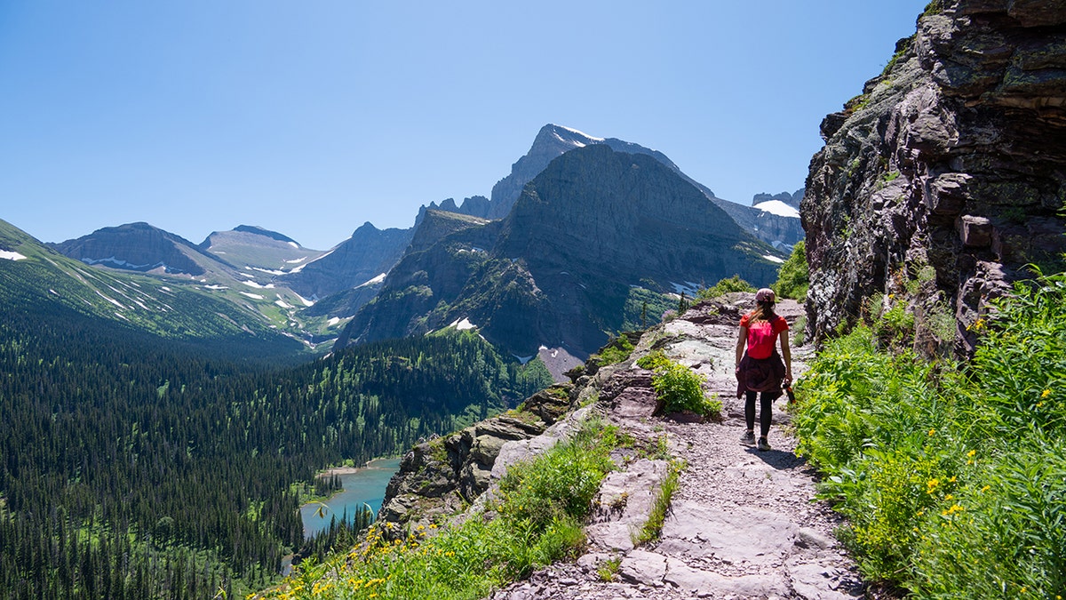 Hiker walking along a narrow mountain trail overlooking a turquoise alpine lake and snow-capped peaks.