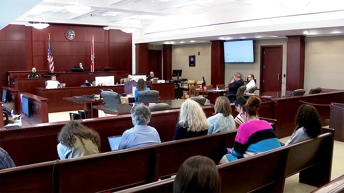 James Melady siedzi na sali sądowej jako sędzia, a inni siedzą