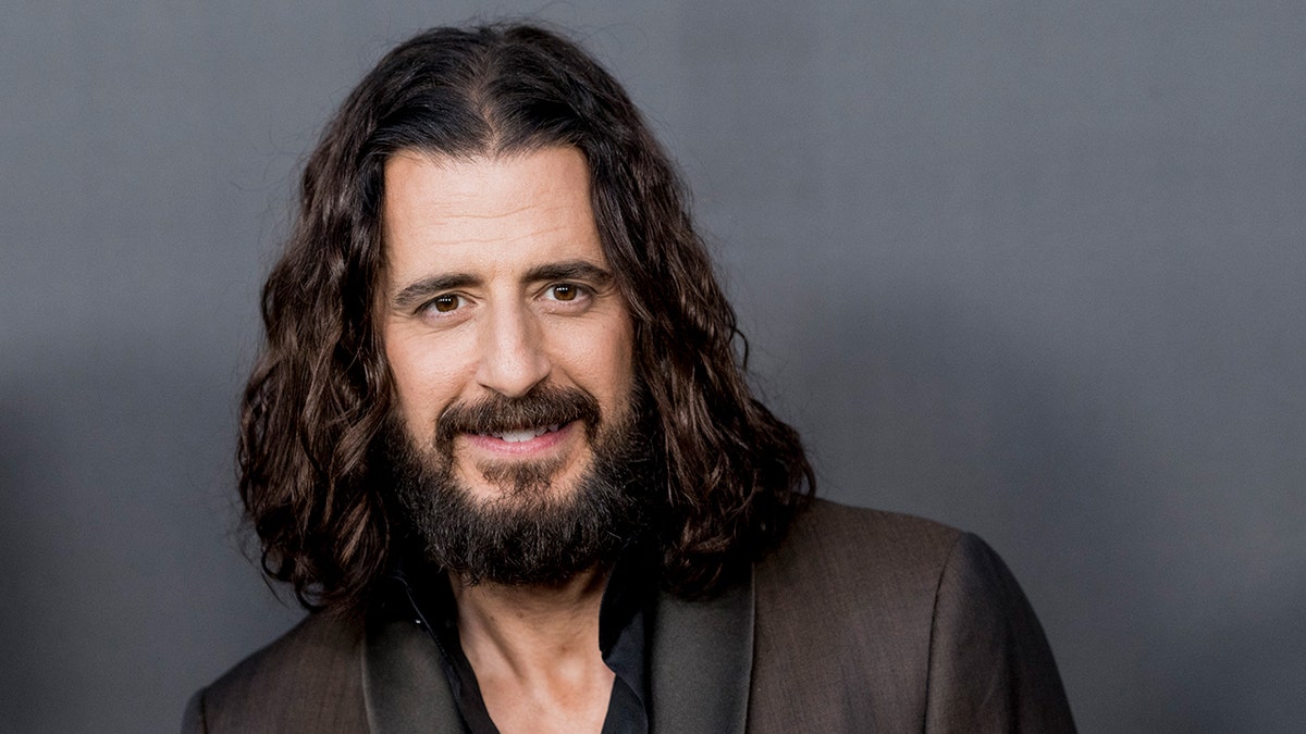 Jonathan Roumie wearing a black shirt and textured gray blazer.